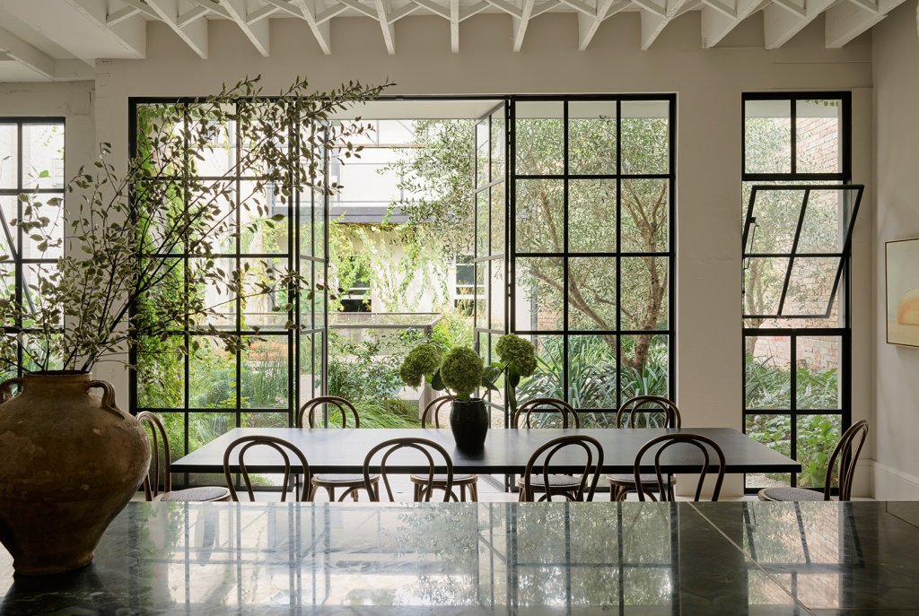 This hidden gem, featured in Belle Magazine, was built by Pamment Projects Building Company in Newtown. The image showcases 10 Tonet timber chairs around a dining table, with a striking black-framed window. The outdoor area features a garden with Olive trees planted to create intimacy and contrast. Designed by The Garden Social, the space is dreamy yet precise.